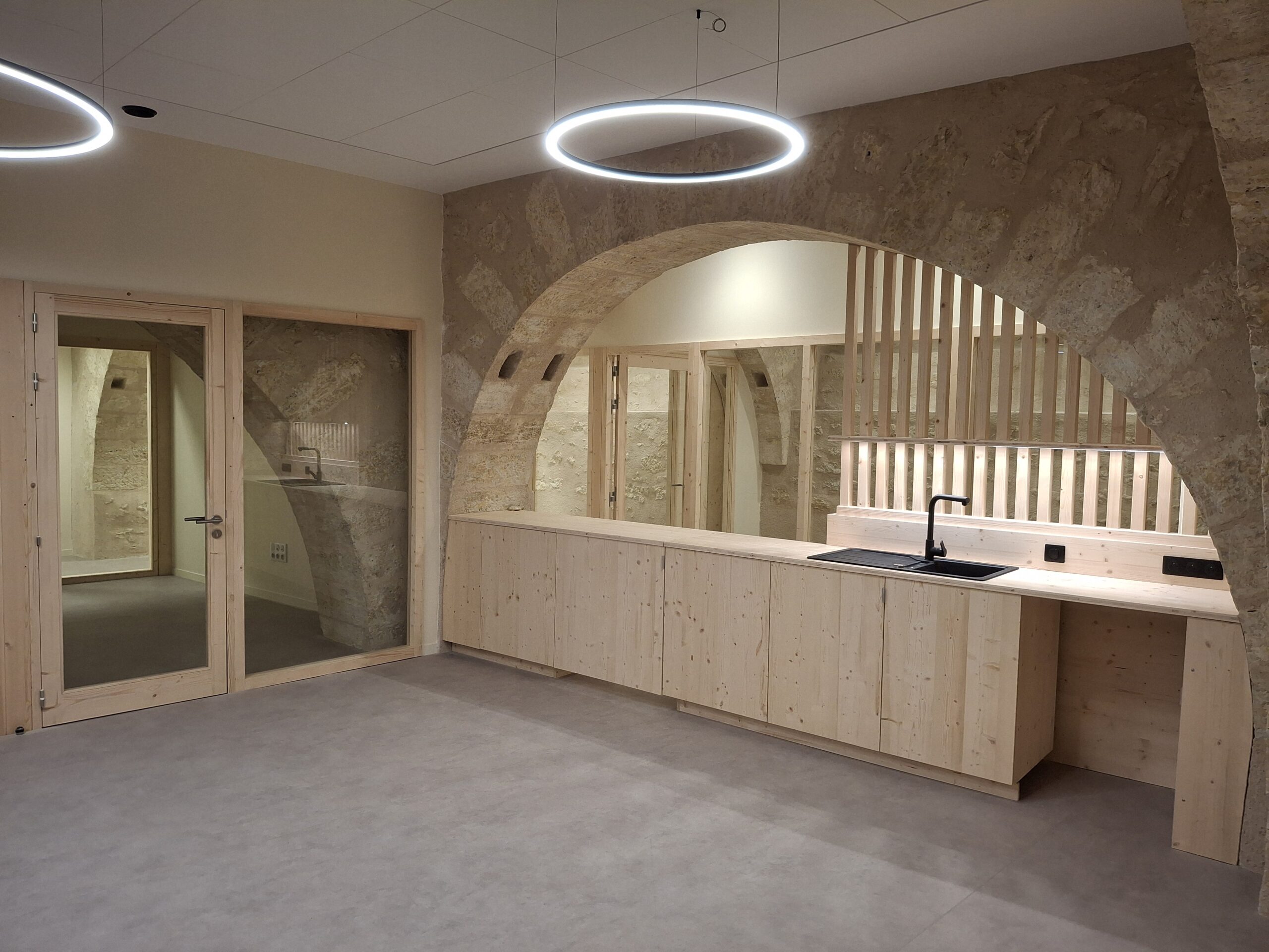 Cuisine minimaliste en pierre et bois clair réalisée par Menuiserie MDB à Occitanie sous grande arche lumineuse,
